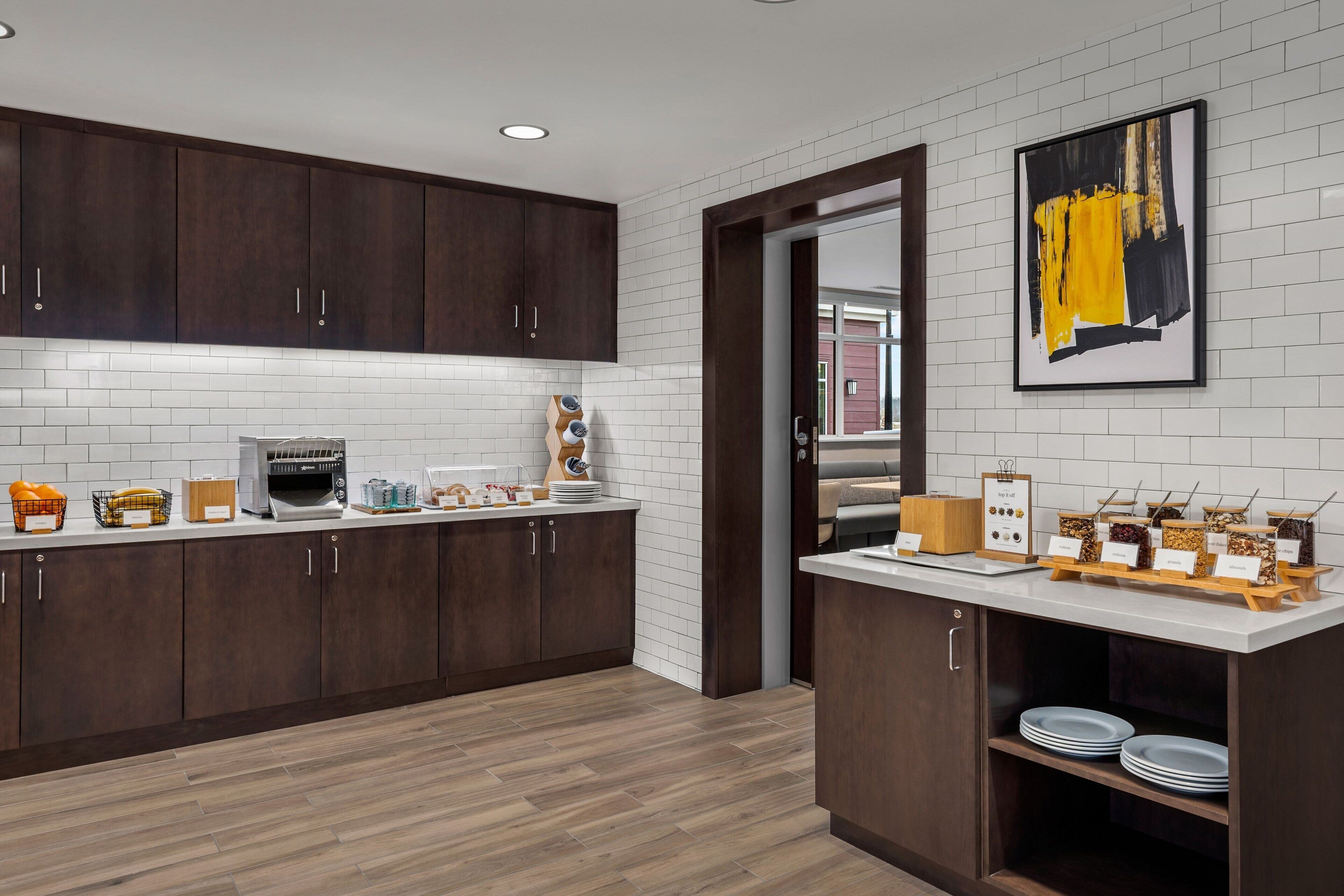 A modern kitchen with dark wooden cabinets, white subway tiles, and a white countertop. There are various kitchen appliances and a buffet setup with labeled containers.