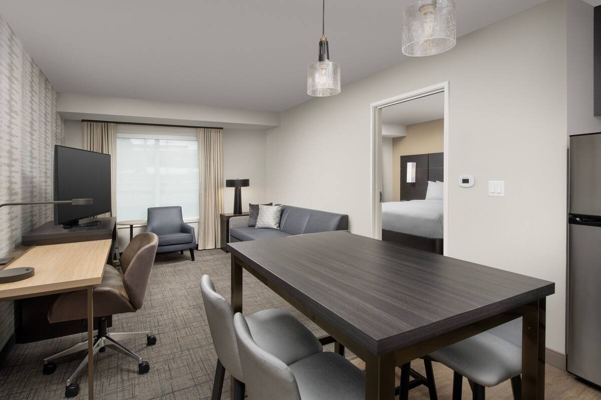Une chambre d’hôtel moderne avec une table à manger en bois, des chaises à revêtement gris, un téléviseur à écran plat, un canapé et une grande fenêtre avec des rideaux.