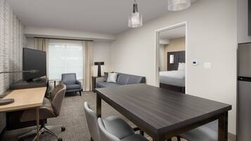 A modern hotel room with a wooden dining table, grey upholstered chairs, a flat-screen TV, a sofa, and a large window with curtains.