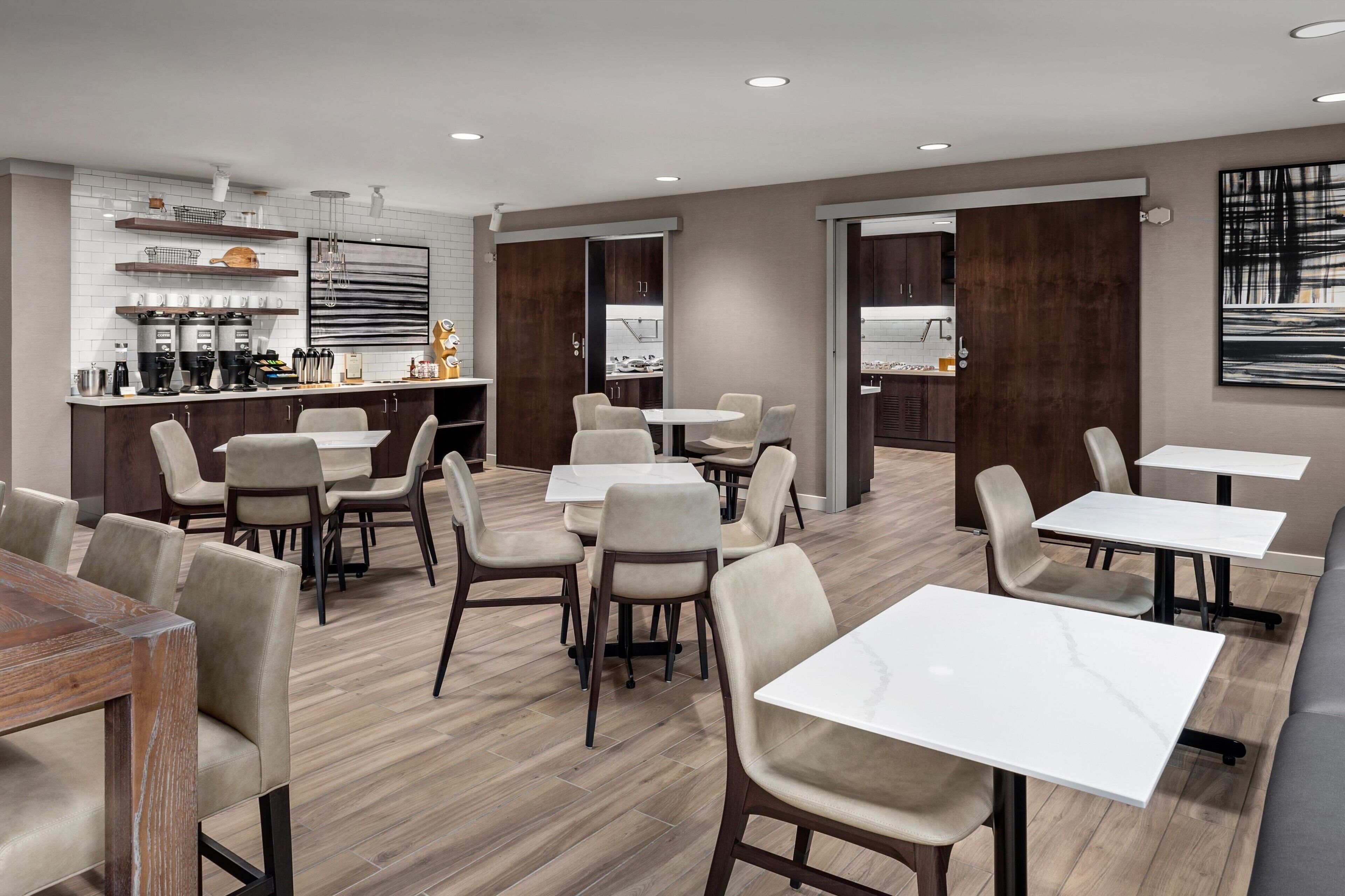 A modern dining area with multiple tables and chairs, a kitchen area with appliances and shelves, and a large abstract painting on the wall.