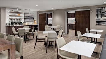 A modern dining area with multiple tables and chairs, a kitchen area with appliances and shelves, and a large abstract painting on the wall.