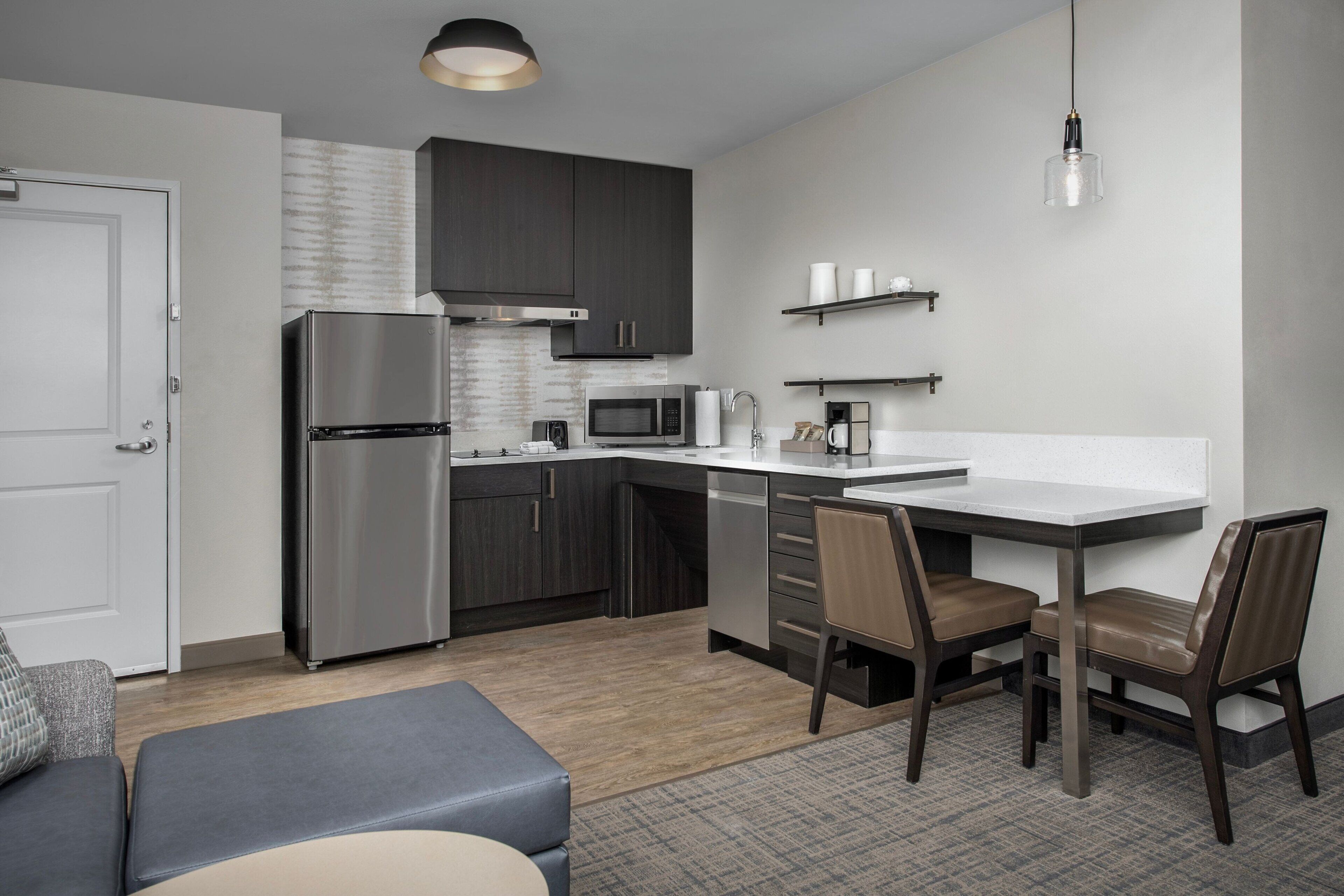 A modern kitchen with dark cabinets, stainless steel appliances, and a white countertop. There is a dining area with two chairs and a small table.