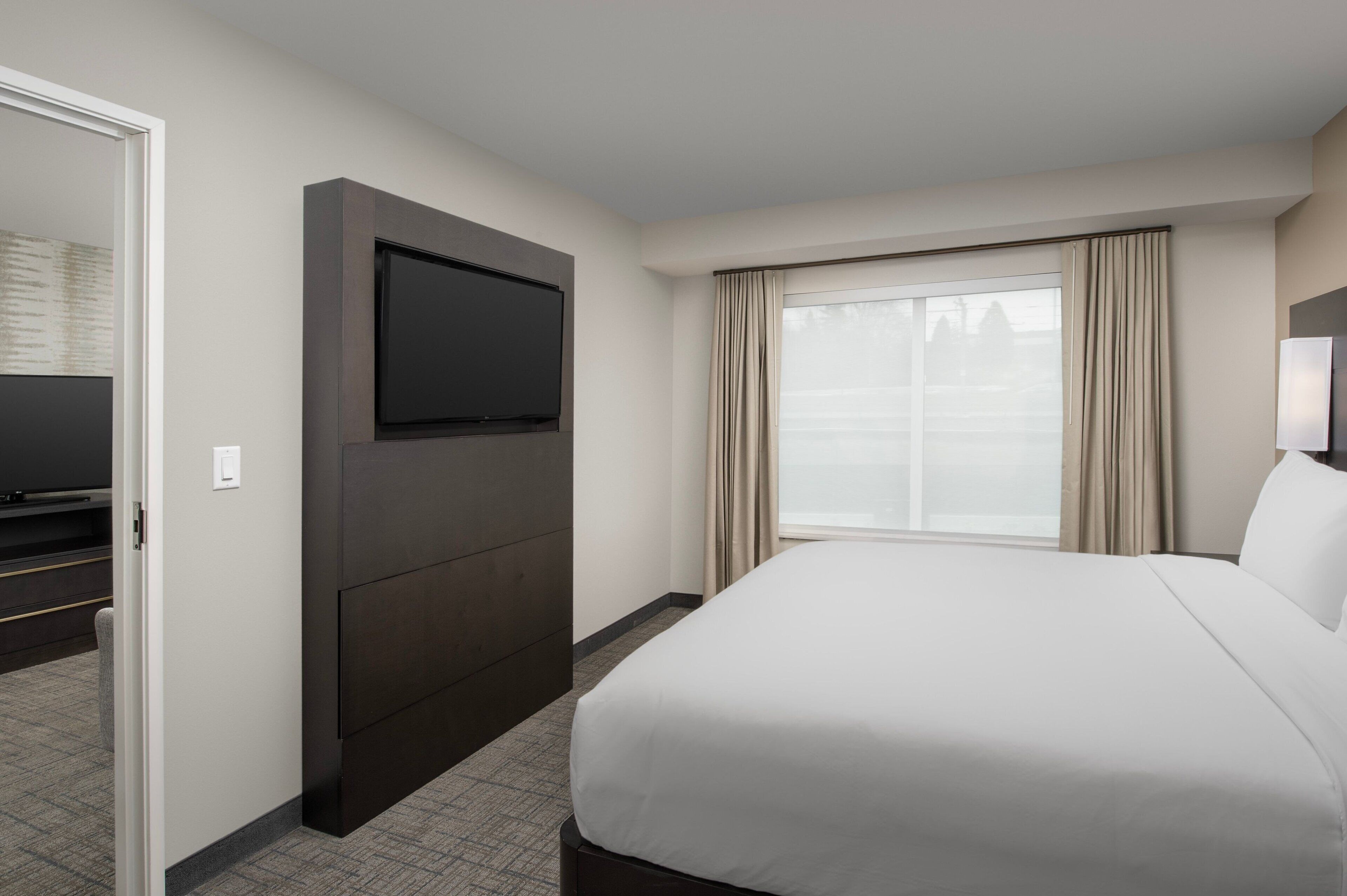 A hotel room with a large bed, a flat-screen TV mounted on the wall, and a large window with curtains.