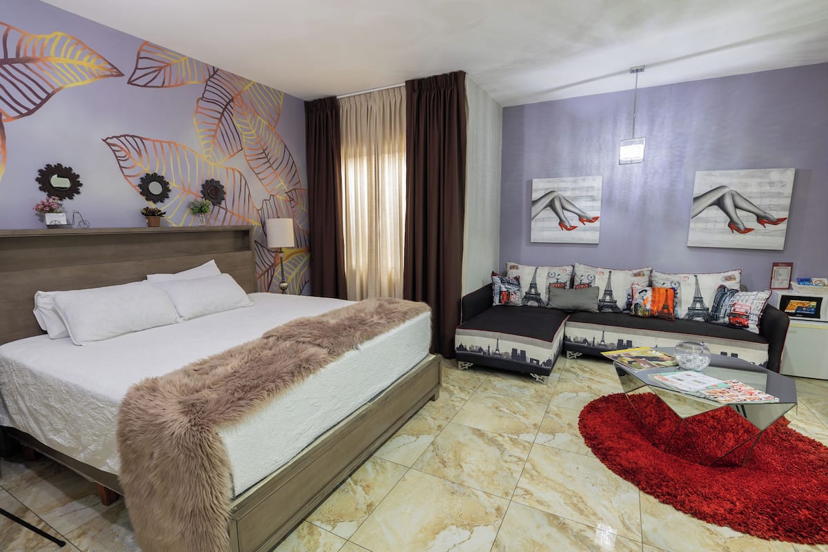 A hotel room with a bed, a sofa, a glass coffee table, and a red rug.