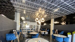 A modern interior with round tables and blue chairs, a patterned ceiling, and a bar area in the background. - Basha Hotel & Restaurant (San Pedro Sula)