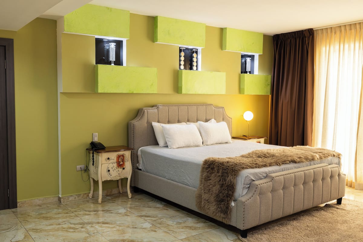 A bedroom with a large bed, a nightstand, a wall-mounted shelf, and a window with curtains.