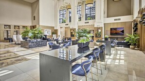 Eine geräumige Lobby mit Barbereich in der Mitte, verschiedenen Sitzgelegenheiten und großen Fenstern für viel Tageslicht.