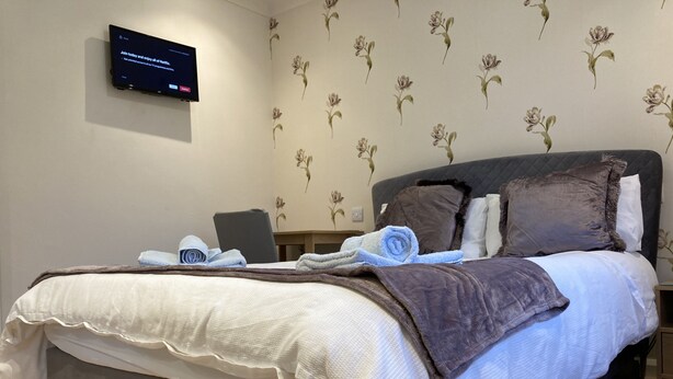 A neatly made bed with a white and brown duvet, a laptop on a bedside table, and a TV mounted on the wall displaying a message. - Spacious Detached Bungalow With Ample Parking (Grantham)