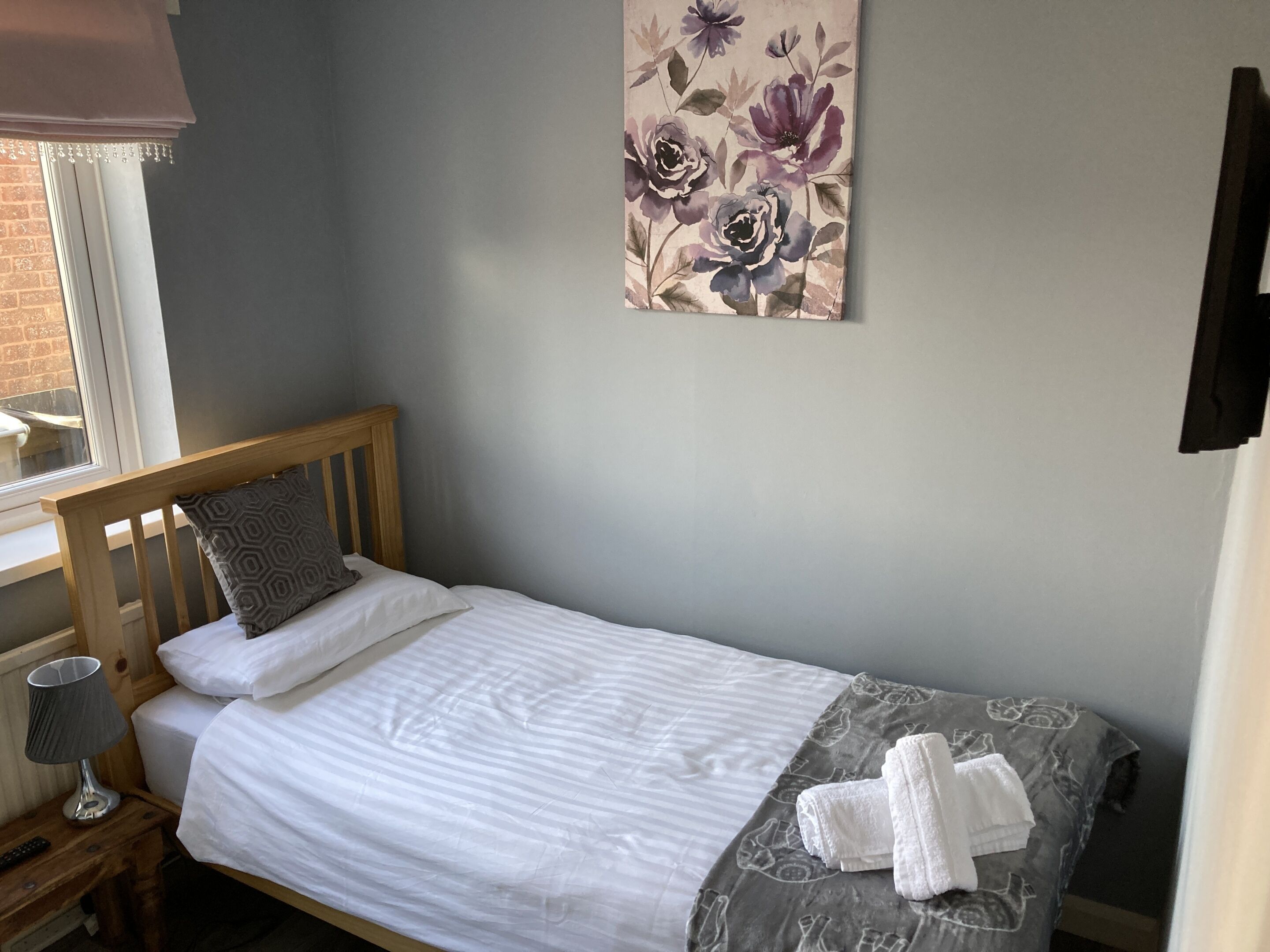 A single bed with a wooden headboard, a grey patterned blanket, and a floral artwork on the wall.