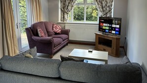 A living room with a purple sofa, a grey sofa, a wooden TV stand, and a window with floral curtains. - Spacious Detached Bungalow With Ample Parking (Grantham)