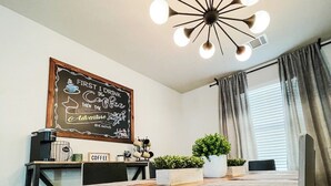 A modern dining area with a wooden table, a chalkboard with a coffee-themed message, a coffee machine, and a potted plant.