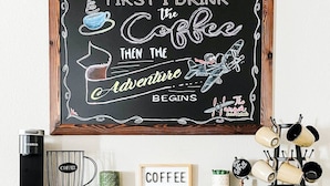 A chalkboard with motivational coffee-related text and illustrations, a coffee maker, a mug, and a coffee-themed sign on a wooden shelf.
