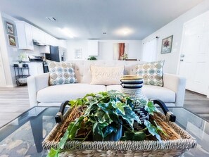 Una sala de estar con una mesa de centro de vidrio, un sofá blanco con cojines estampados, y una cocina al fondo.