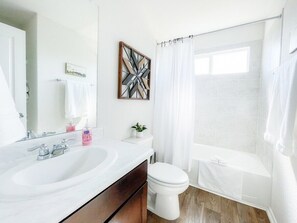 Ein modernes Badezimmer mit einem weißen Waschbecken, einer Toilette und einer Badewanne. Über dem Waschbecken befindet sich ein Spiegel, an der Wand ein gerahmtes Kunstwerk und auf der Ablagefläche ein kleiner Topfpflanze.