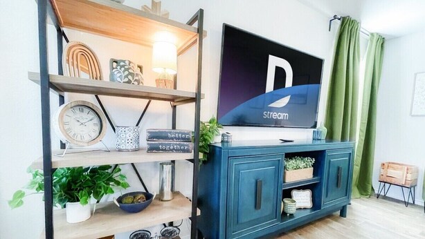 A living room with a TV mounted on the wall, a blue cabinet, and shelves with decorative items. - Stylish Family Home  Near Lackland  POOL! (San Antonio)