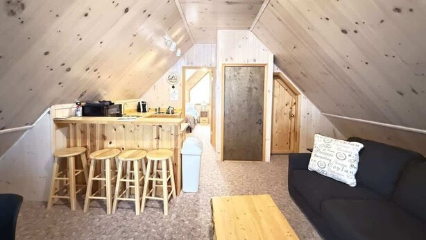 A cozy attic room with a kitchenette, wooden stools, a sofa, and a small table.