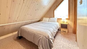 A bedroom with a sloped ceiling, a bed with a beige comforter, a nightstand with a lamp, and a window providing natural light.
