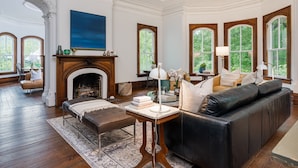 A spacious living room with a fireplace, leather sofa, and large windows offering a view of greenery. - Magnolia House estate - family reunions, micro-weddings, retreats. (Hot Springs)