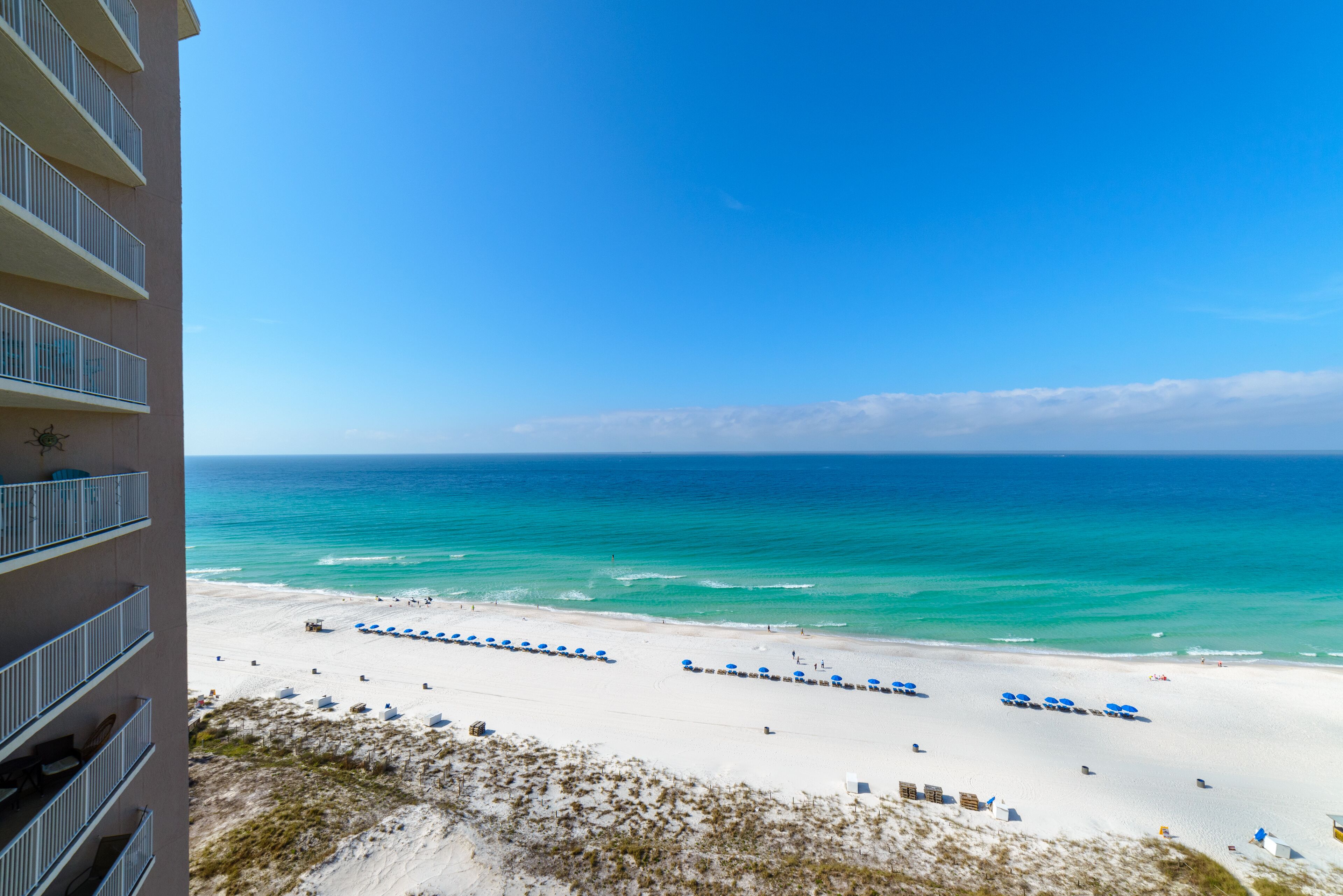 Blick auf einen Strand vom Balkon aus, mit weißem Sand, klarem, blauem Wasser und wolkenlosem Himmel.