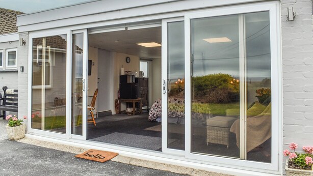 Un bâtiment moderne doté de grandes portes vitrées, d’un espace cuisine visible et d’un petit coin salon extérieur avec un fauteuil en osier et un banc.