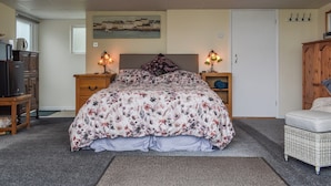 Une chambre à coucher avec un lit, deux tables de chevet avec des lampes, une armoire en bois, un fauteuil en osier et un tableau accroché au mur.