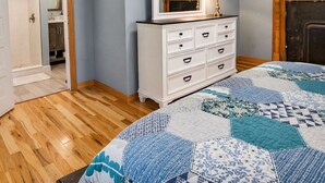 Une chambre avec un plancher en bois, une commode blanche, un miroir, un lit recouvert d’une couette bleue et blanche, et une cheminée.