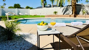 A poolside area with a table holding a bowl of fruit and a glass of orange juice, lounge chairs, and a palm tree.