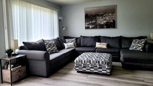 Ein modernes Wohnzimmer mit einem Ecksofa, einem gemusterten Hocker und einem Stadtbild an der Wand.