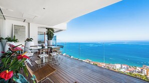 A balcony with a view of the sea, a dining area with chairs and a table, and potted plants.