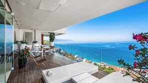A modern outdoor balcony with a view of the sea, furnished with white sofas and a table.