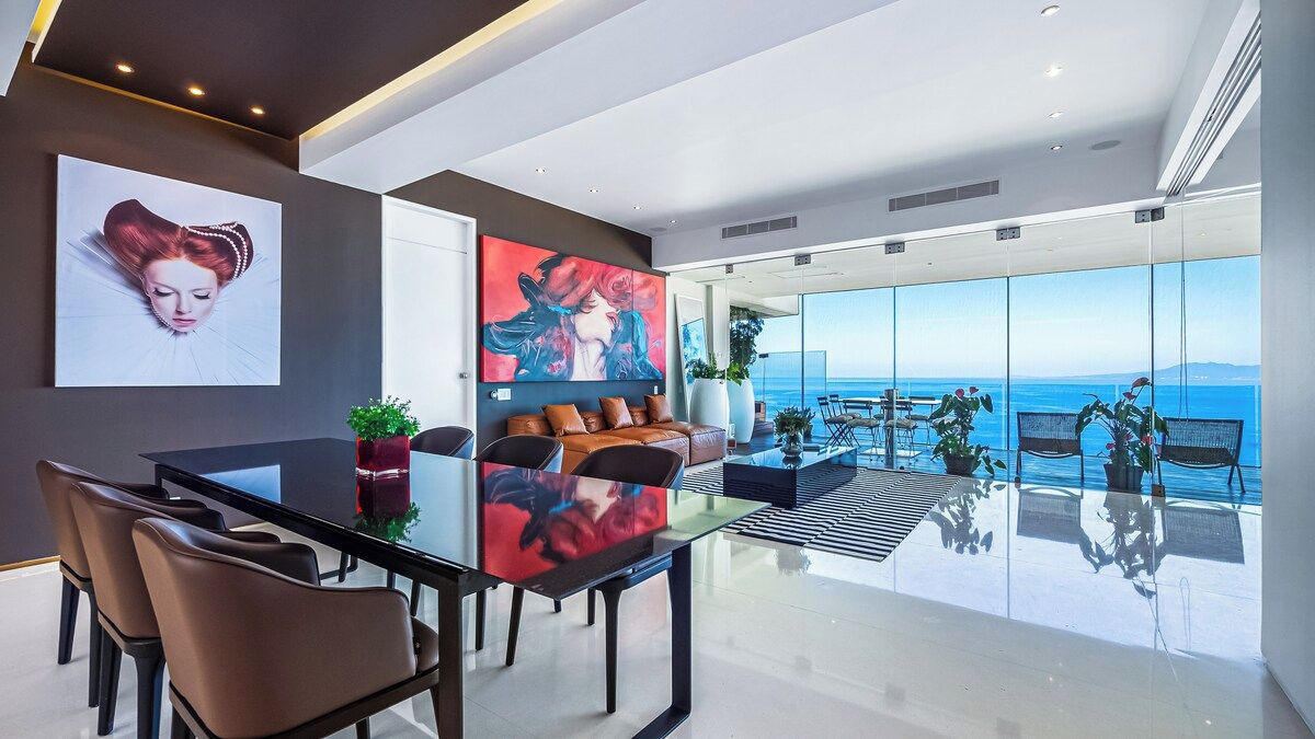 A modern interior with a dining area, a sofa, and a large window overlooking the sea.