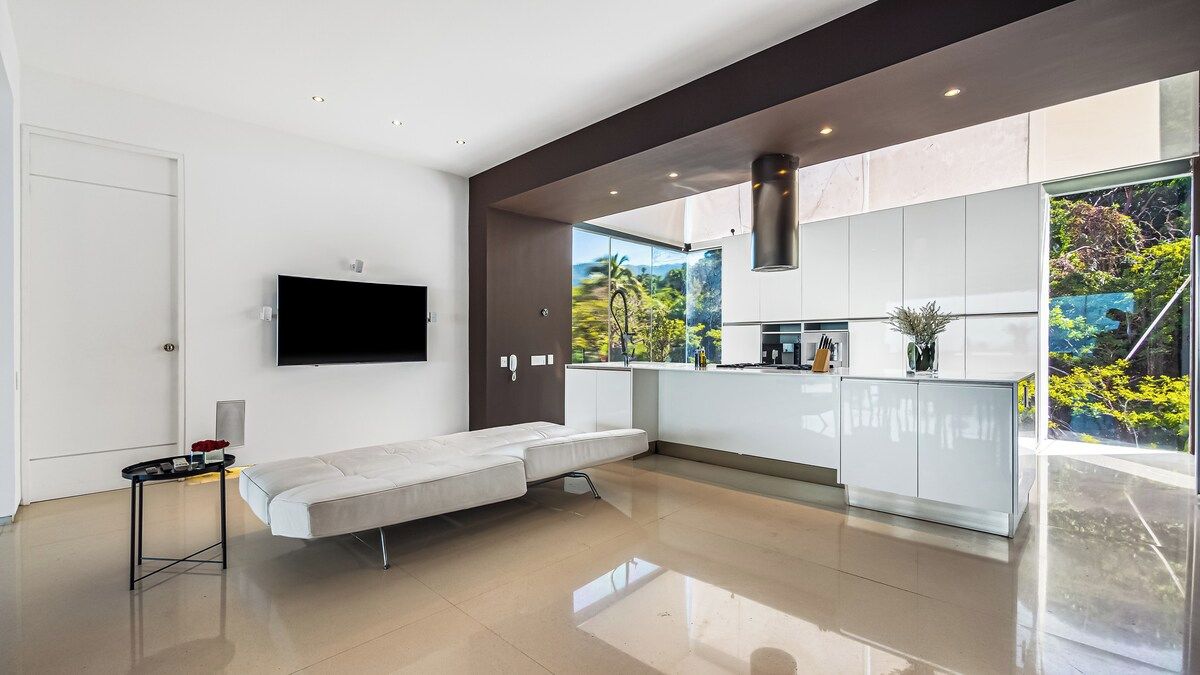 Une cuisine moderne avec des meubles blancs, un téléviseur à écran plat fixé au mur et une porte-fenêtre coulissante donnant sur un balcon avec vue sur la verdure.