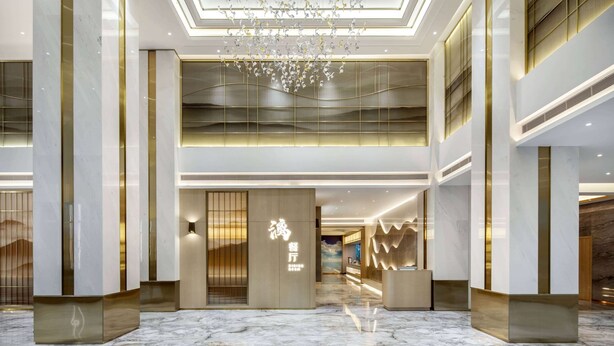 Un vestíbulo de hotel con diseño moderno, que presenta un piso de mármol, un mostrador de recepción y una lámpara decorativa en el techo.