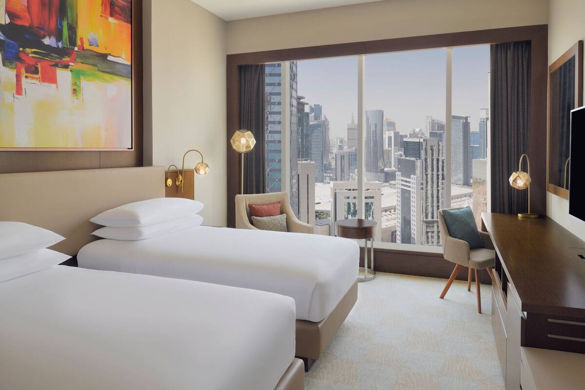 A hotel room with two beds, a desk, a chair, a large window with a city view, and a painting on the wall.