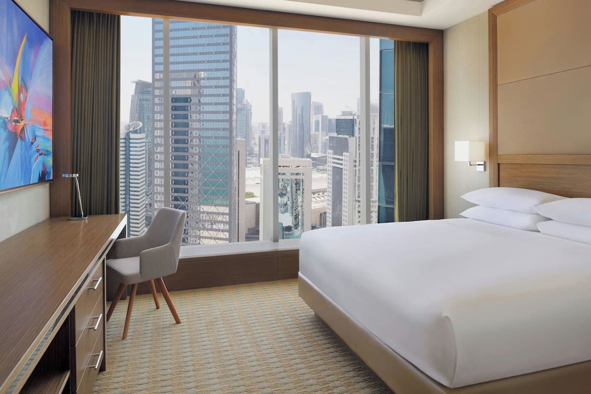 A hotel room with a bed, a desk, a chair, and a view of a cityscape through large windows.