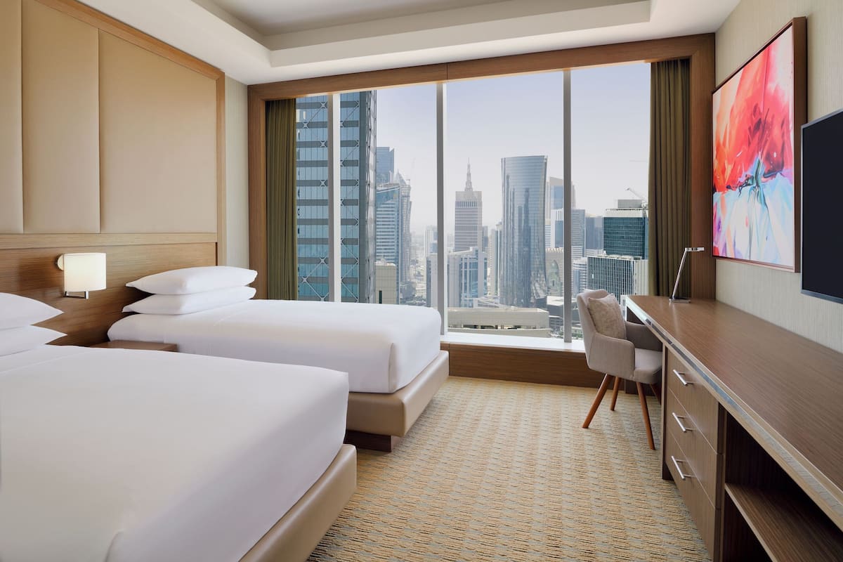 A hotel room with two beds, a desk, and a large window offering a city view.
