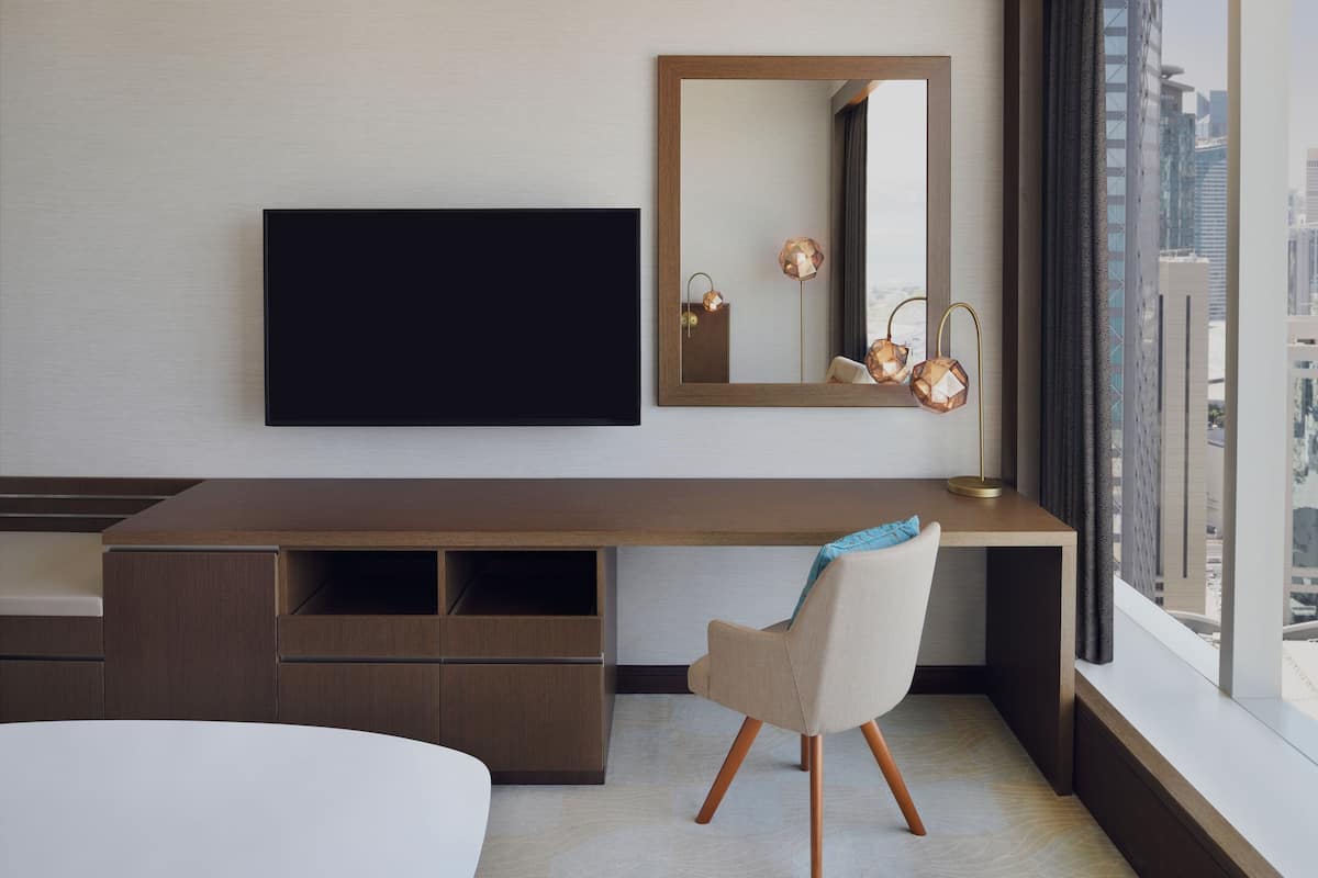 A modern hotel room with a large desk, a flat-screen TV, a chair, and a large window offering a city view.
