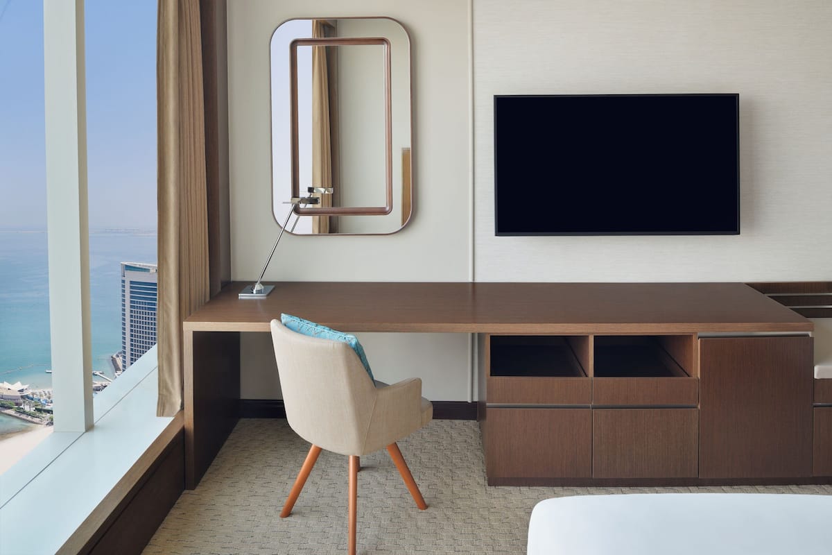 A modern hotel room with a large desk, a flat-screen TV, a chair, and a view of the city and ocean.
