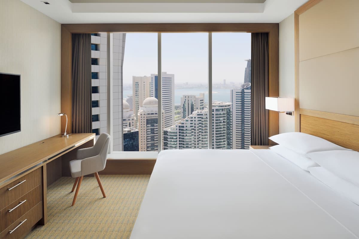 A hotel room with a large bed, a desk with a chair, a TV, and a view of a cityscape through a large window.