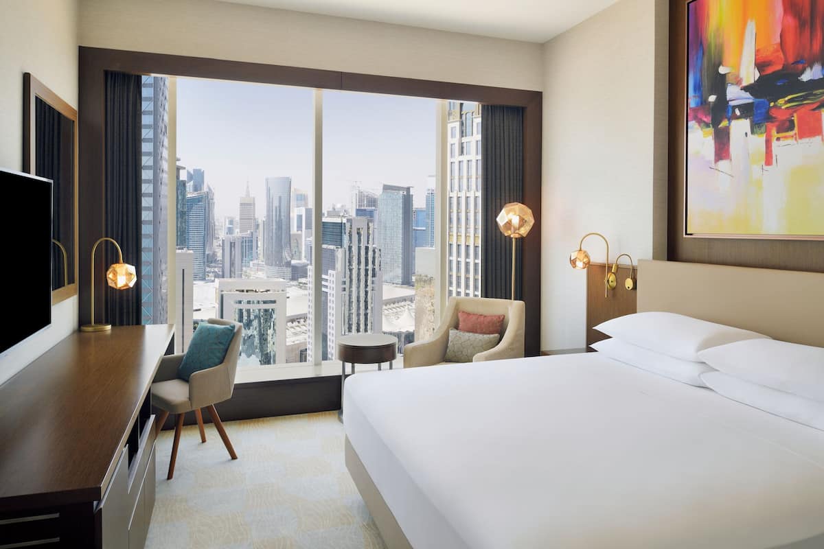 A hotel room with a large bed, a desk, a chair, a small table, and a view of a cityscape through large windows.