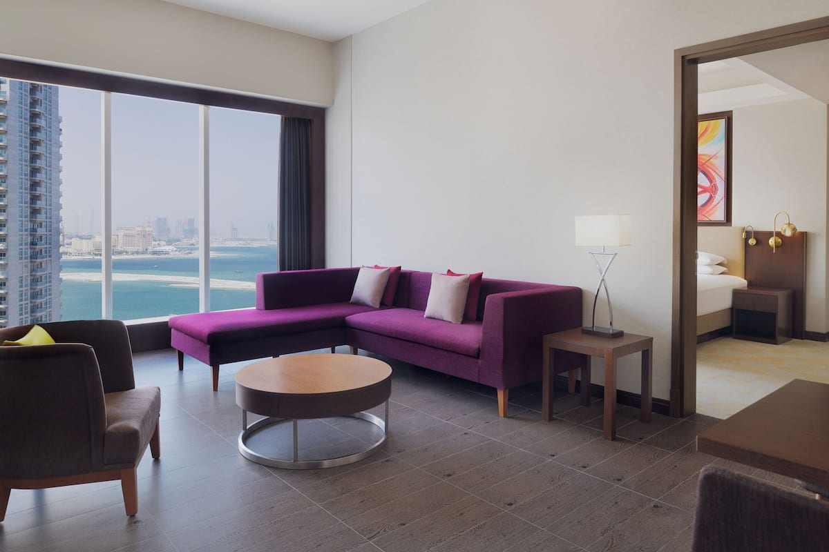 A modern hotel room with a large window offering a city view, a purple sectional sofa, a round coffee table, and a small side table.