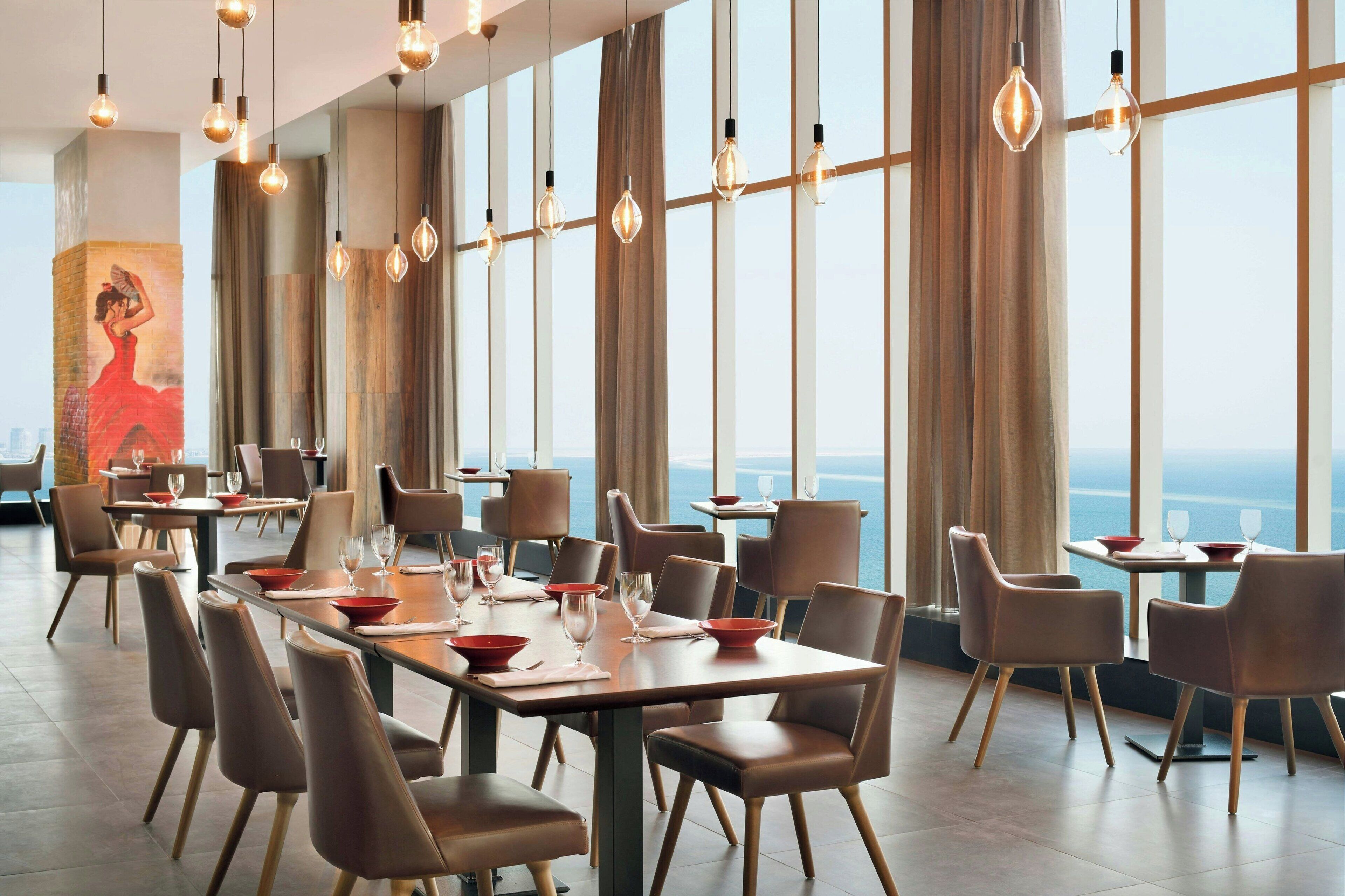 Un restaurant avec des tables dressées pour le service, de grandes fenêtres et des luminaires suspendus.