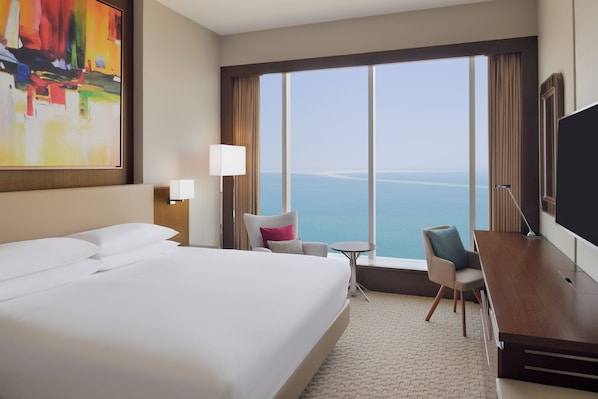 Ein Hotelzimmer mit einem großen Bett, einem Schreibtisch, einem Sessel, einem kleinen Tisch und Meerblick.