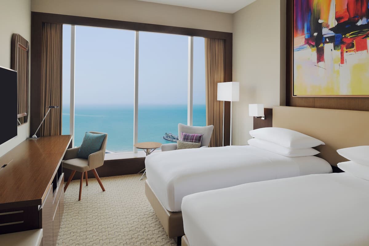 A hotel room with a large window, a desk, a chair, two beds, and a painting on the wall.