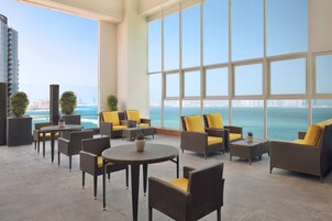 Un salón moderno con mobiliario de mimbre y cojines amarillos, grandes ventanas que ofrecen vistas al mar y al horizonte de la ciudad.