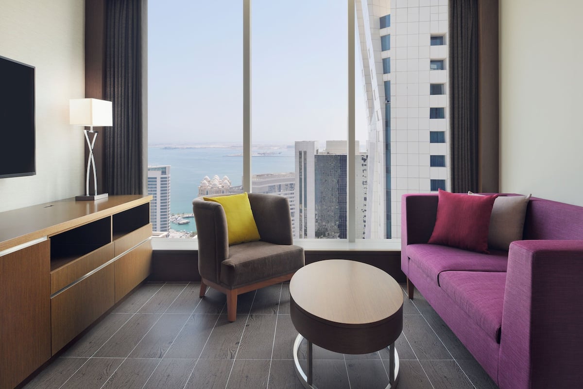 A modern hotel room with a large window offering a city view, a sofa, a chair, a small round table, and a flat-screen TV.