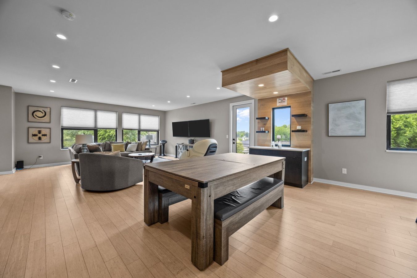 A spacious living room with a wooden dining table, a gray sofa, and a large window offering a view of greenery.