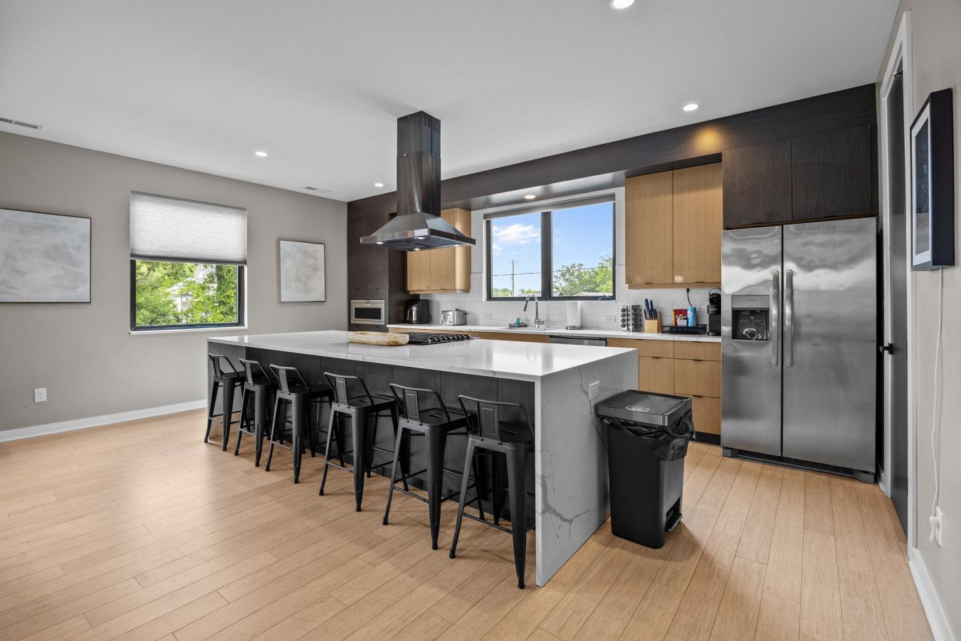 A modern kitchen with a central island, bar stools, stainless steel appliances, and a large window providing natural light.