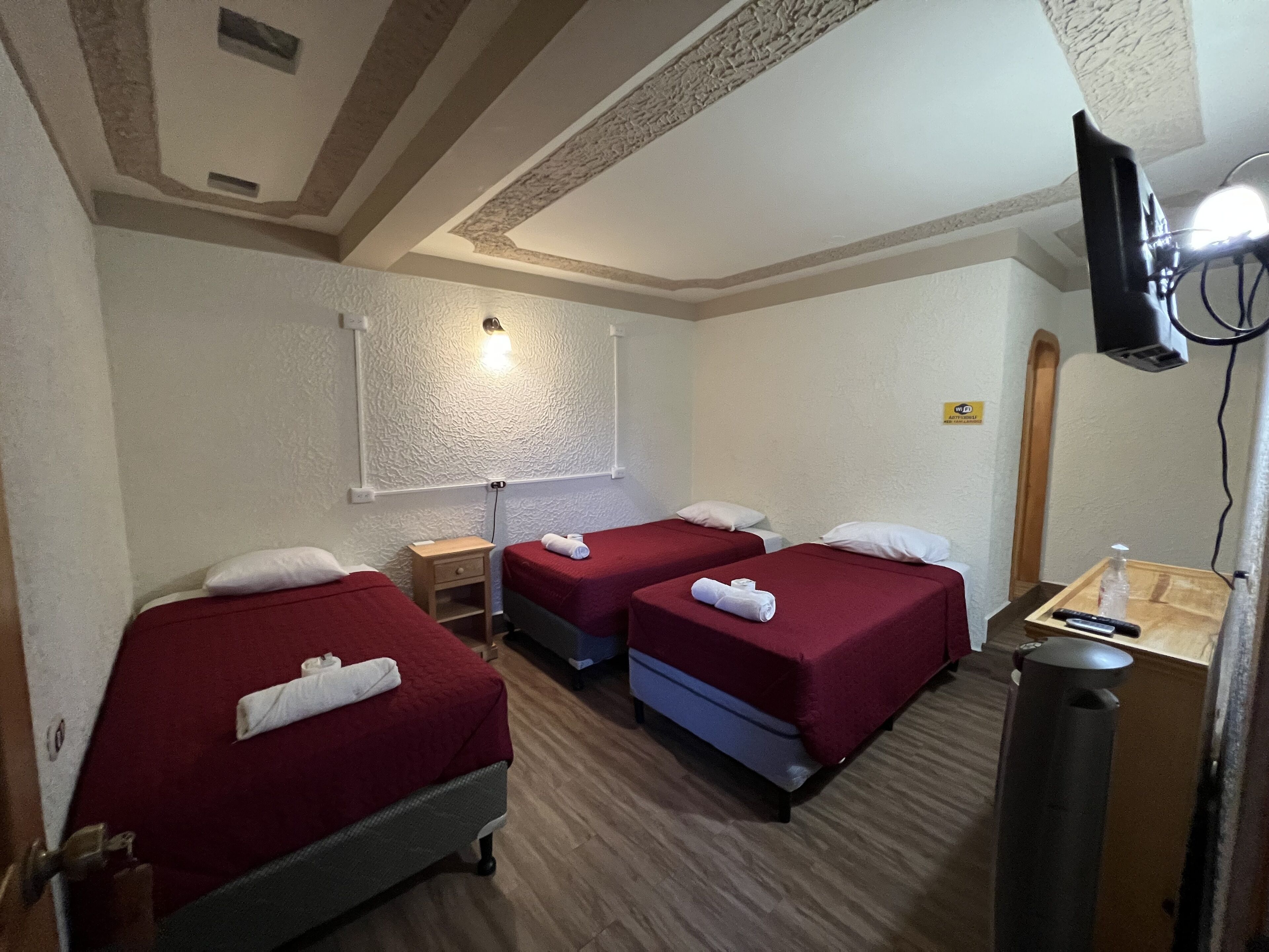 Camera d'albergo con tre letti singoli, una TV fissata al muro, un tavolino con una bottiglia e un telecomando, e una luce a soffitto.