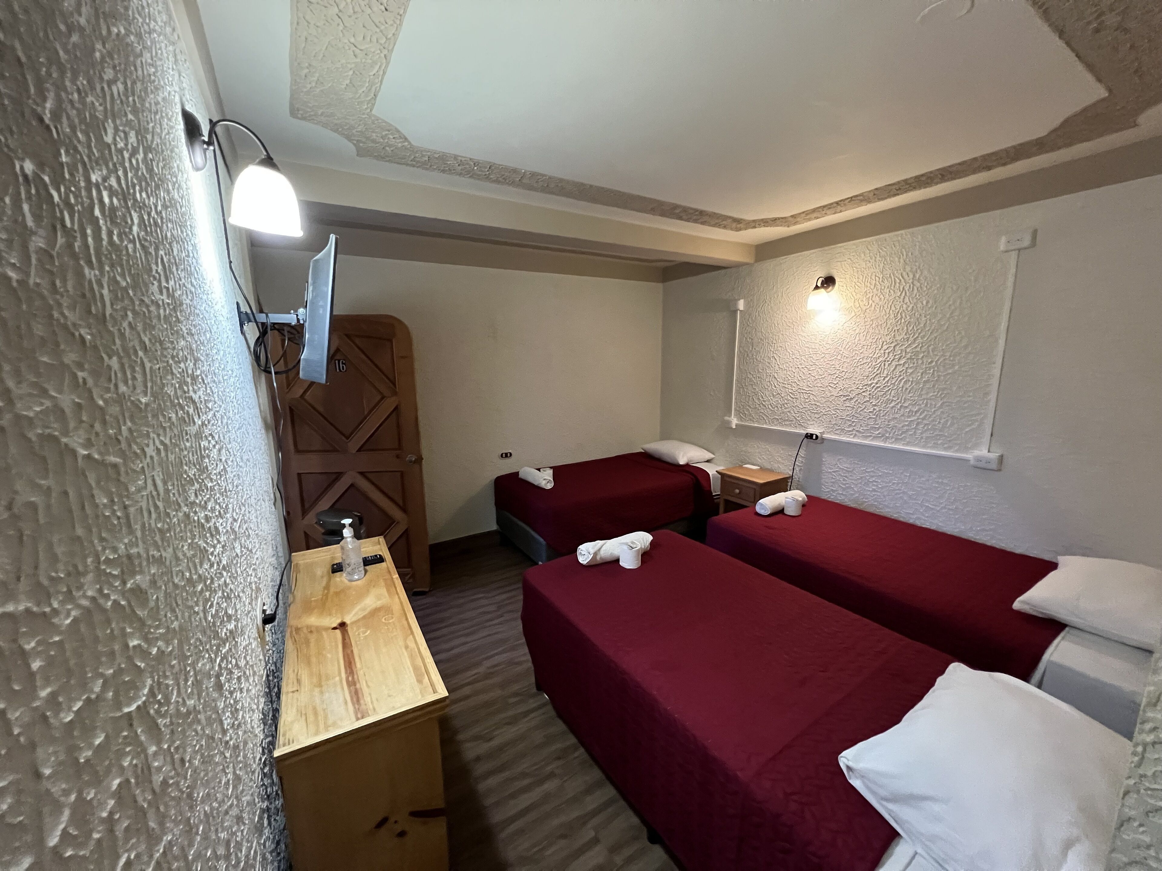 A small hotel room with two single beds, a wooden nightstand, a small table with a bottle and tissues, and a wall-mounted lamp.